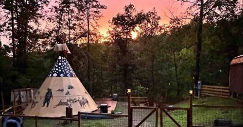 A colorful teepee stands in a wooded area at sunset, surrounded by trees and a fenced yard.