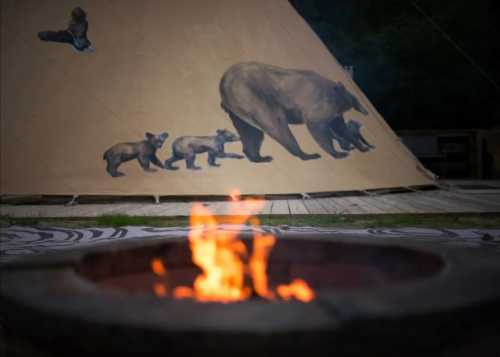 A campfire in the foreground with a teepee featuring bear murals in the background.