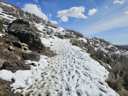 A snowy trail winds through a rocky landscape under a blue sky with scattered clouds.