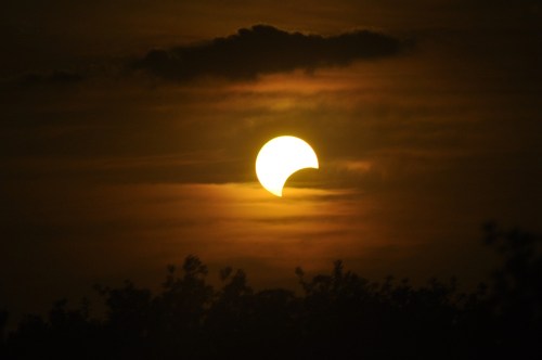 A partial solar eclipse with a bright sun partially obscured by clouds, set against a darkening sky.