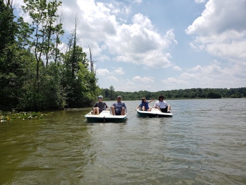 Three people relax on paddle boats in a calm lake surrounded by trees and a blue sky with fluffy clouds.