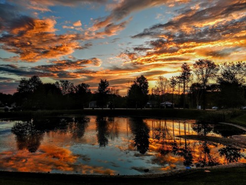 A serene sunset over a pond, with vibrant clouds reflecting in the water and trees silhouetted against the sky.