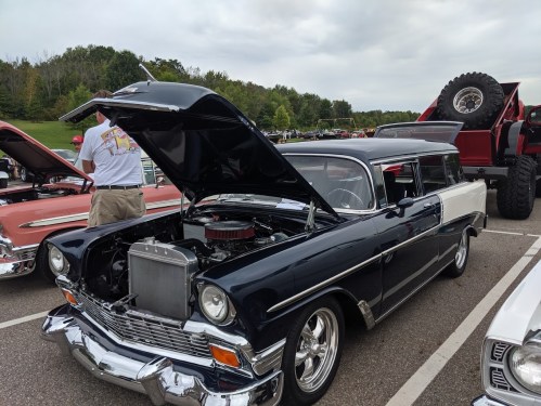 A classic black and white 1956 car with the hood open, showcasing its engine, at a car show with other vehicles in the background.