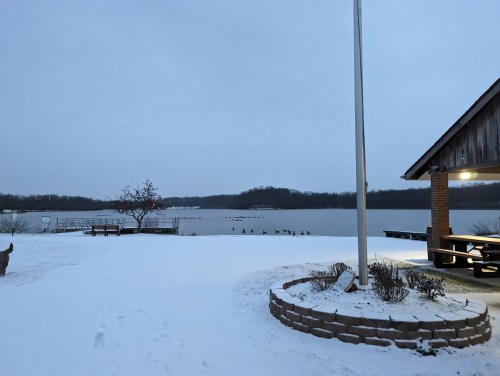 A snowy lakeside scene at dusk, featuring a pavilion, a flagpole, and ducks on the water in the background.