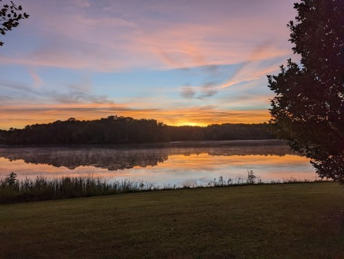 A serene lake at sunrise, reflecting colorful skies and surrounded by trees and grassy shorelines.
