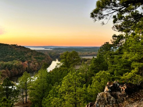 A scenic view of a river winding through lush green trees at sunset, with a colorful sky in the background.