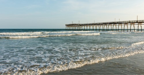 A serene beach scene featuring gentle waves and a wooden pier extending into the calm ocean under a clear sky.