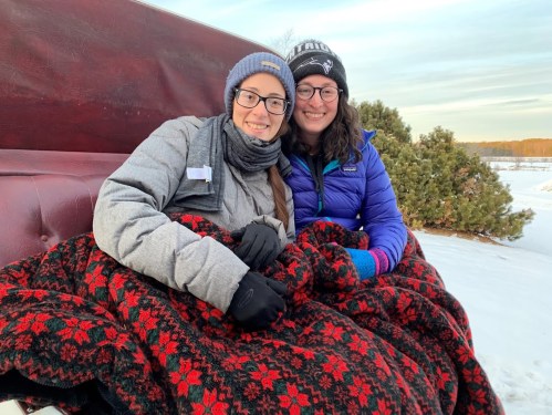 Two women sit together on a sleigh, wrapped in a red and black blanket, smiling in a snowy landscape.