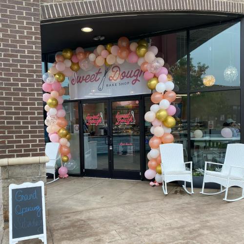 Entrance of Sweet Dough Bake Shop adorned with a colorful balloon arch and a sign for the grand opening. White rocking chairs outside.