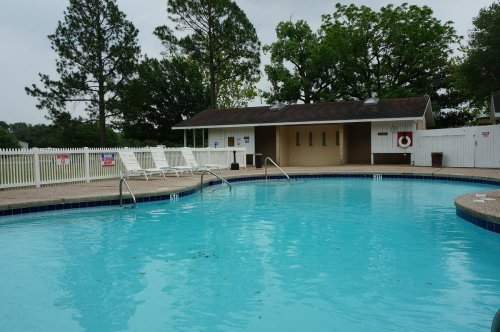 A clear swimming pool with lounge chairs nearby, surrounded by trees and a small building in the background.