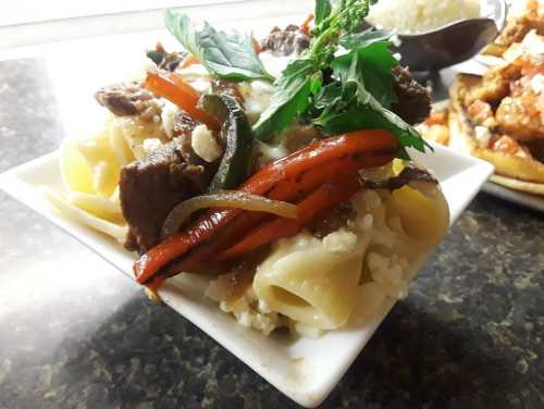 A bowl of pasta topped with beef, colorful bell peppers, and fresh herbs on a dark countertop.