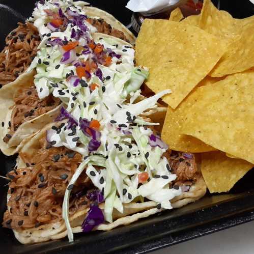 Three tacos filled with shredded meat, topped with cabbage and colorful veggies, served with tortilla chips on the side.