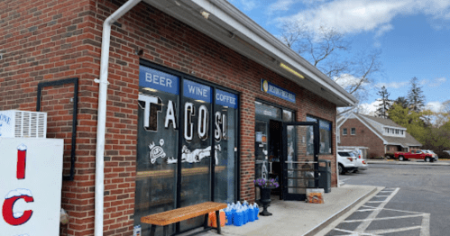 A brick storefront with signs for tacos, beer, wine, and coffee, featuring a bench outside and a parking lot.