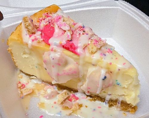 A slice of cheesecake topped with colorful frosting, sprinkles, and bits of cookie, served in a white takeout container.