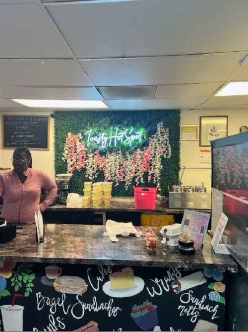 A café counter with a neon sign, greenery backdrop, and a staff member preparing food. Colorful menu items displayed.