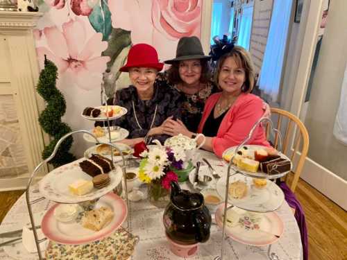 Three women in stylish hats enjoy tea and desserts at a beautifully set table with flowers and a floral backdrop.