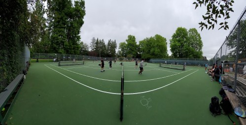 A panoramic view of a tennis court with players in action and spectators seated along the sidelines under cloudy skies.