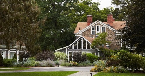 A charming stone house with a sloped roof, surrounded by lush greenery and colorful plants in a serene setting.