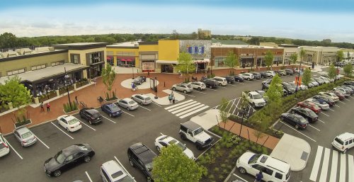 Aerial view of a shopping center with parked cars, storefronts, and pedestrians walking along a crosswalk.