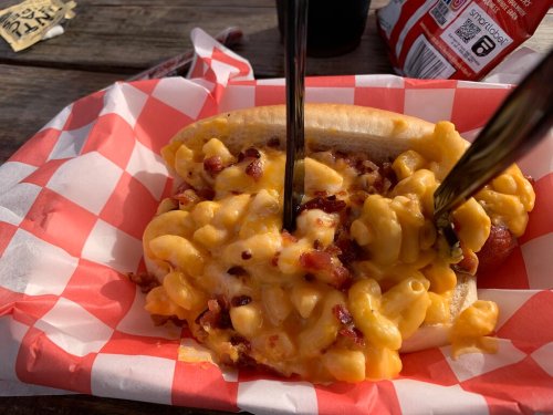 A hot dog topped with macaroni and cheese and bacon bits, served in a red and white checkered paper.