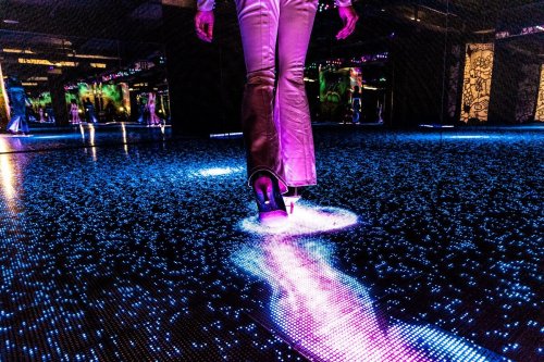 A person in shiny pants walks on a glowing, illuminated floor with colorful lights reflecting around them.