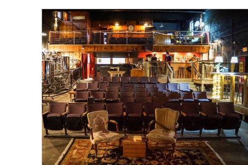 A cozy theater interior with empty seats, a stage, and two vintage chairs in the foreground. Warm lighting creates an inviting atmosphere.