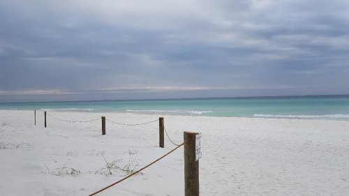 A serene beach scene with white sand, gentle waves, and a cloudy sky, marked by wooden posts and rope fencing.