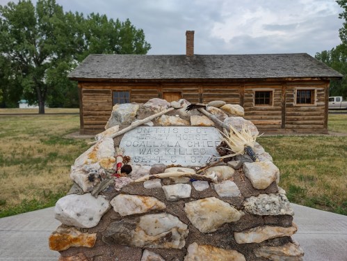 Memorial stone marking the spot where Crazy Horse was killed, with a log cabin in the background.