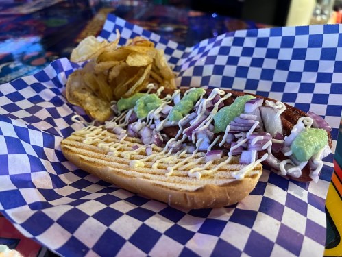A hot dog topped with green relish and onions, served with potato chips on a checkered paper plate.
