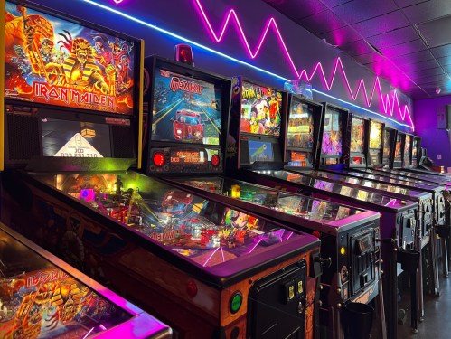 A row of colorful pinball machines illuminated by neon lights in a vibrant arcade setting.
