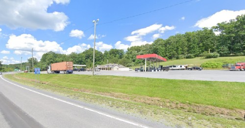 A gas station with trucks parked nearby, set against a backdrop of green hills and a blue sky with clouds.
