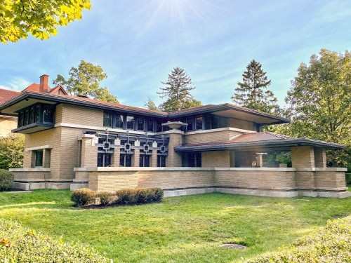 A modernist building with a flat roof, large windows, and a landscaped lawn, surrounded by trees under a clear blue sky.