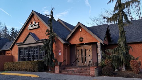 Exterior of a restaurant with a rustic orange facade, large wooden doors, and evergreen trees flanking the entrance.