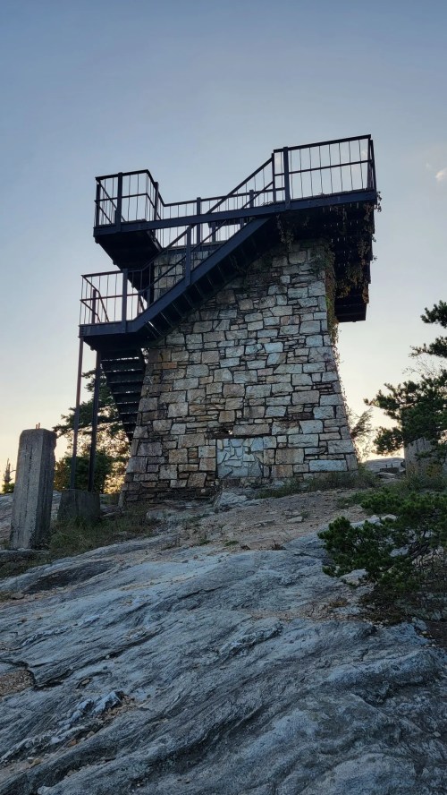 A stone observation tower with a metal staircase, surrounded by rocky terrain and trees under a clear sky.