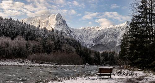 A snowy landscape featuring a river, a bench, and majestic mountains under a partly cloudy sky.