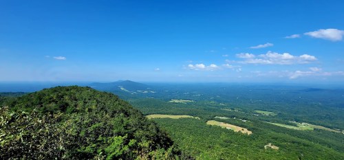 A panoramic view of lush green hills and valleys under a clear blue sky, with distant mountains on the horizon.