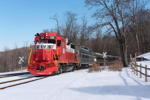 A red train engine with passenger cars travels through a snowy landscape, surrounded by trees and a wooden fence.