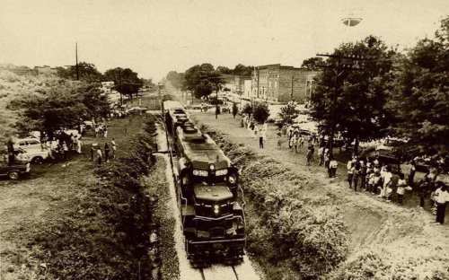 A vintage black-and-white photo of a train passing through a small town, with crowds gathered along the tracks.