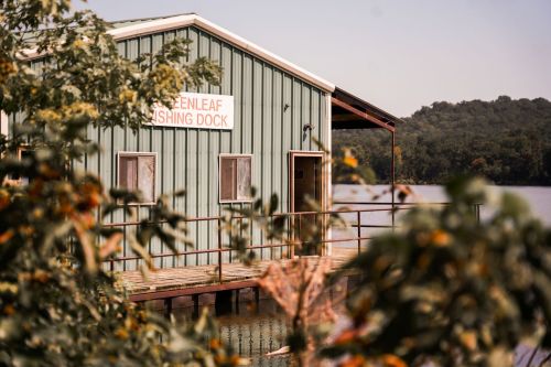 A green fishing dock building by the water, surrounded by trees and a clear sky.