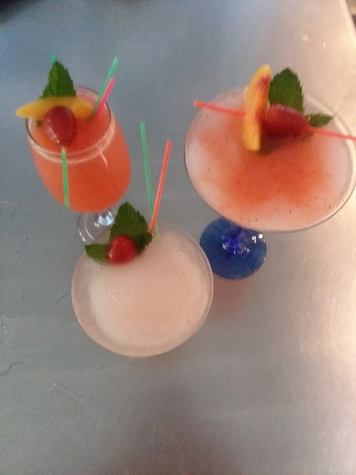 Three colorful cocktails with fruit garnishes and straws, served in various glassware on a metallic surface.
