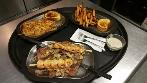 A tray with grilled steak topped with onions and shrimp skewers, sweet potato fries, hash browns, and toasted bread.