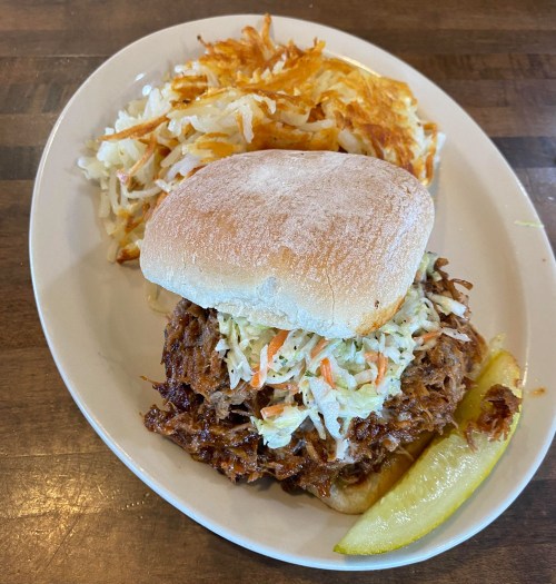 A pulled pork sandwich topped with coleslaw, served with hash browns and a pickle on a white plate.