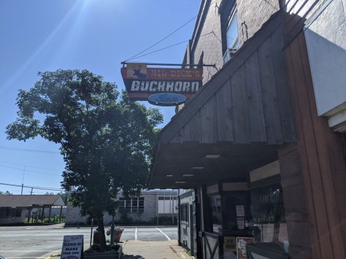 Sign for "Buckhorn" restaurant hangs above a wooden building, with a clear blue sky and tree in the background.