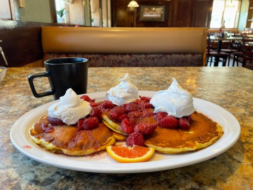 A plate of pancakes topped with raspberries and whipped cream, served with a slice of orange and a coffee cup in the background.