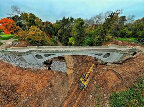 A newly constructed bridge spans a construction site surrounded by trees and autumn foliage. Heavy machinery is visible below.