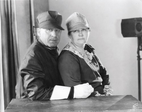 A man in a cap and a woman in a hat sit closely together at a table, both looking directly at the camera.