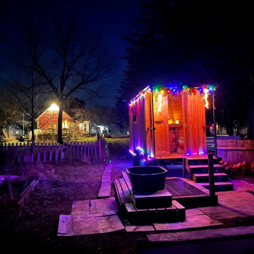A cozy cabin illuminated with colorful lights at night, surrounded by trees and a peaceful outdoor setting.