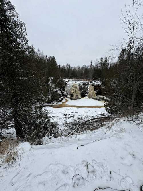 A snowy landscape featuring a waterfall surrounded by trees and a river with brown water. Overcast sky above.