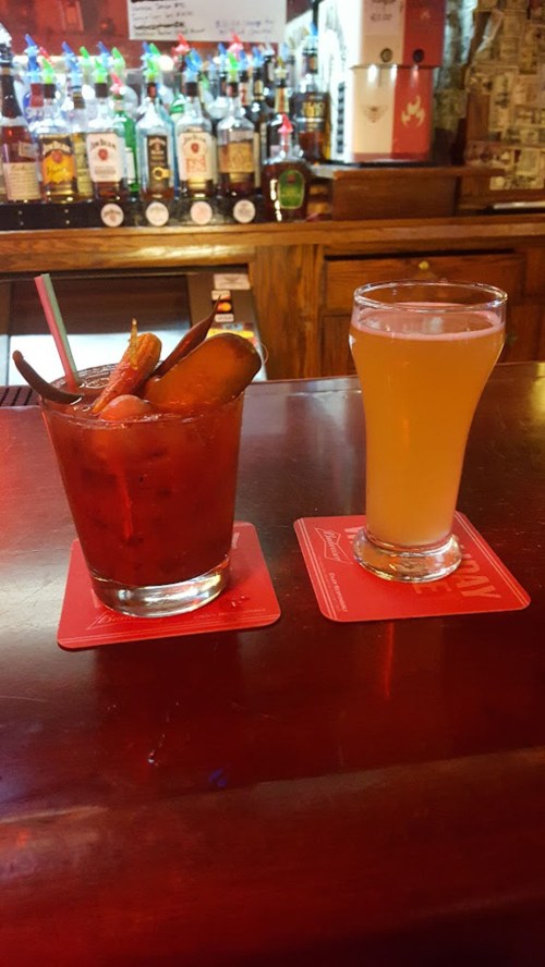 A cocktail with garnishes and a light beer sit on a bar counter, with bottles of liquor in the background.