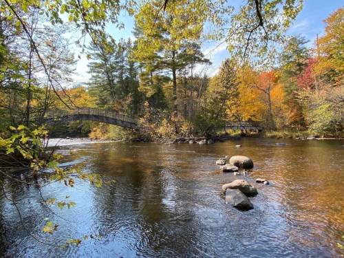 A serene river scene with a wooden bridge, surrounded by colorful autumn foliage and smooth rocks in the water.